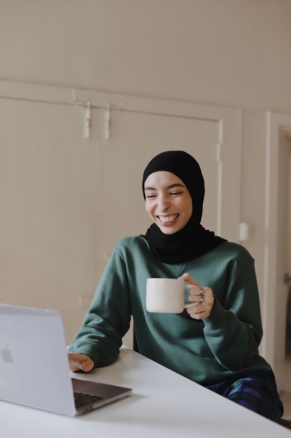 A smiling woman in green hijab holding a white mug using a laptop